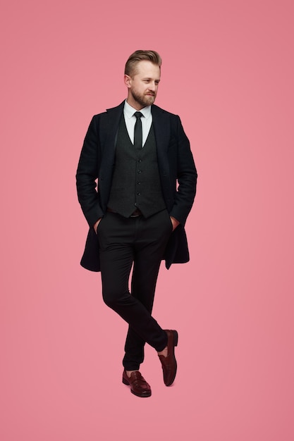 Full body of confident adult bearded gentleman wearing classy black suit with tie keeping hands in pockets while standing against pink background