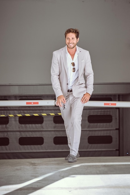 Full body of confident male entrepreneur in elegant suit and tie looking at camera while walking near shelves with crossed legs