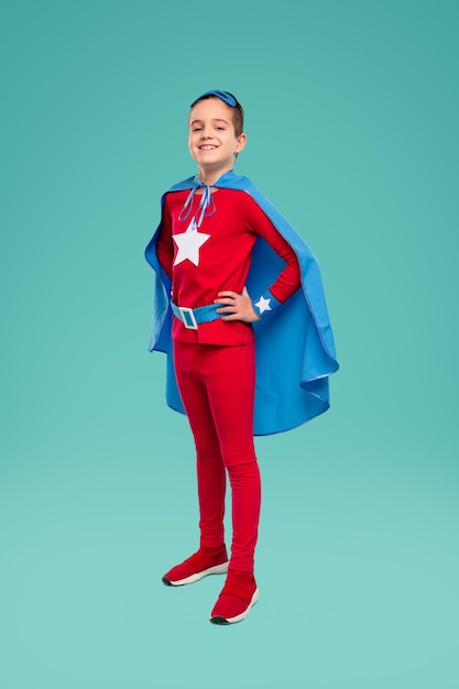 Full body of confident smiling boy in bright red superhero outfit, with blue cape keeping hands on waist and looking at camera against turquoise background