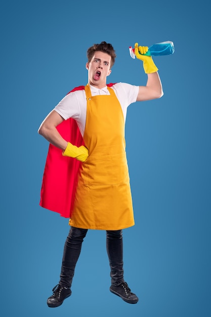 Full body of crazy young male in yellow apron and red superhero cloak holding bottle, with detergent in raised hand and screaming with funny grimace against blue background