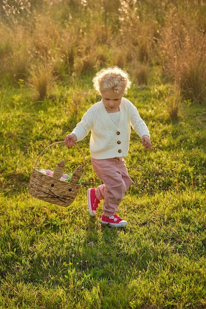 Полнотелая милая маленькая девочка с вьющимися волосами в стильном наряде прогуливается по травянистому зеленому лугу на летней природе и смотрит вниз