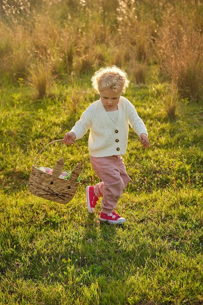 Full body cute little girl with curly hair wearing stylish outfit strolling on grassy verdant meadow in summer nature and looking down