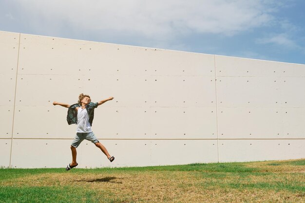 Foto corpo pieno di ragazzo energico in abiti casual che salta fuori terra con le braccia alzate sul prato verde contro il muro