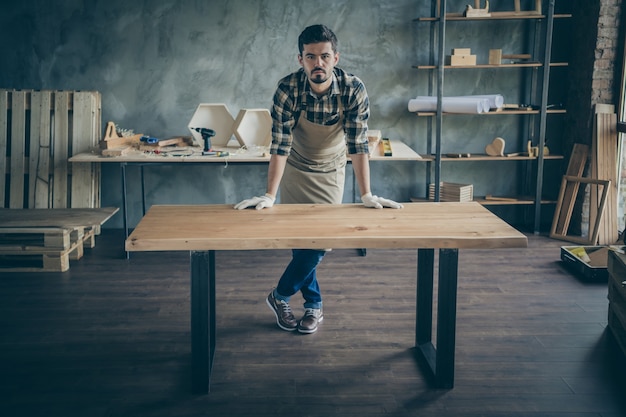 Full body  handsome guy master leaning hands on handmade slab table advertising good work selling website own wooden business industry woodwork shop garage indoors