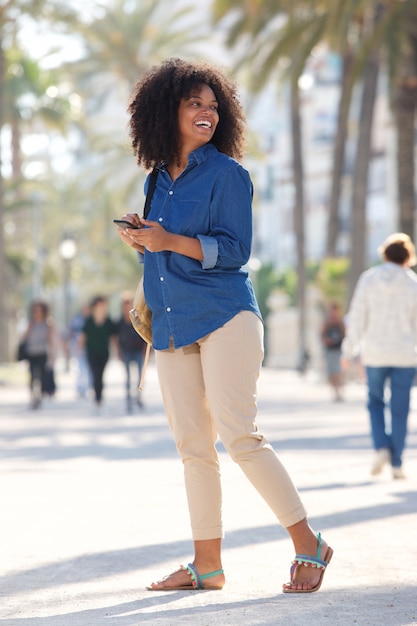 Full body happy woman walking outside with smart phone