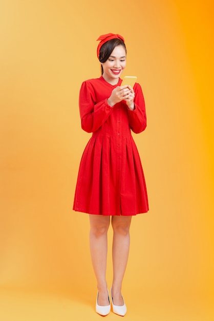 Full body of happy young Asian woman using phone, wearing red dress while standing over colorful background.