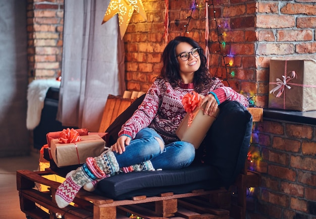 Full body image of brunette female in eyeglasses dressed in a jeans and a red sweater posing on a wooden sofa in a room with Christmas decoration.