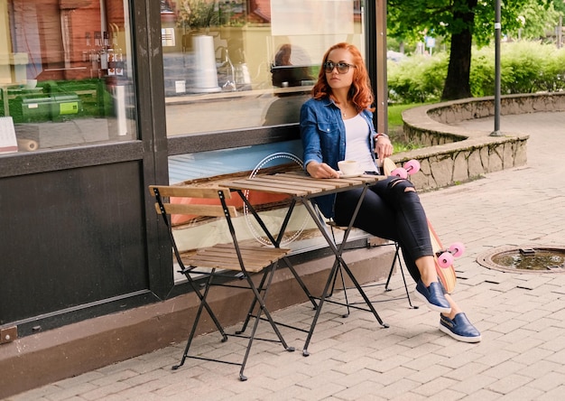Full body imgae of redhead female drinks coffee at the table in a cafe on a street.