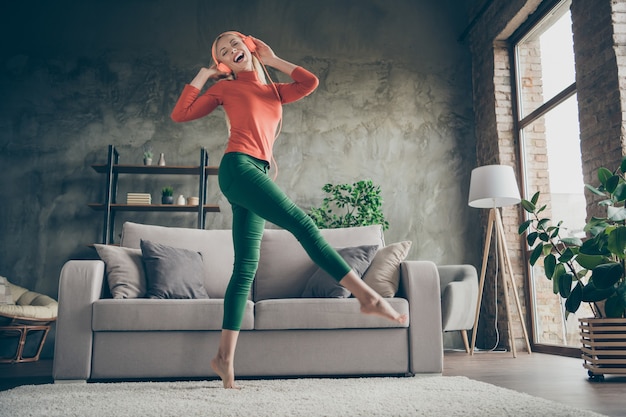 Full body lage hoek weergave foto van vrij grappige dame bedrijf telefoon luisteren lied moderne oranje koptelefoon dansen in de buurt van bank casual outfit woonkamer binnenshuis