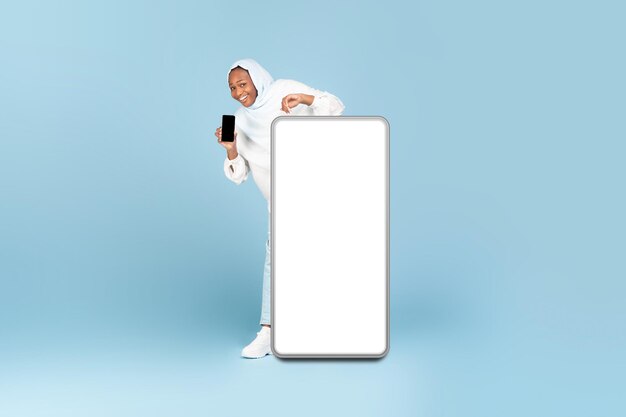 Full body length of black muslim woman peeking out standing behind big smartphone with white blank screen mockup