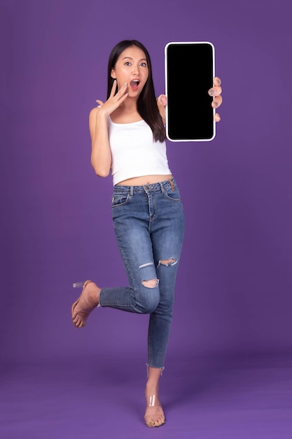Full body length photo portrait of beautiful asian young woman\
excited surprised girl showing big smart phone with blank screen\
white screen isolated on purple background mock up image