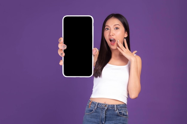 美しいアジアの若い女性の全身写真の肖像画紫色の背景に分離された空白の画面の白い画面で大きなスマートフォンを示す興奮した驚きの女の子モックアップ画像