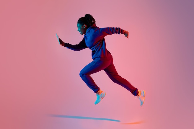 Full body length shot of modern sporty lady jumping or running using cellphone over pink neon background side view