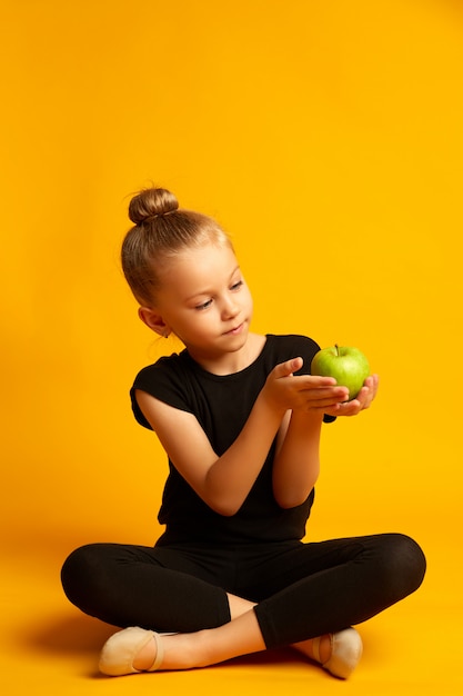 Full body little girl in leotard looking at green apple while sitting against yellow background during break in training