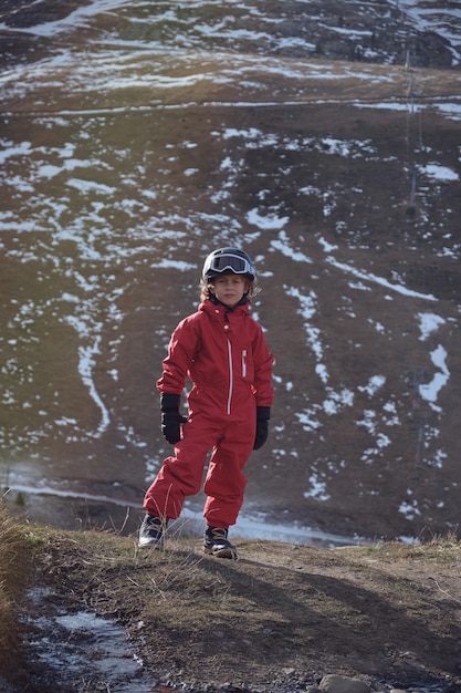 Full body little traveler in outerwear and helmet with goggles looking at camera while standing on slope on snowy mountain on ski resort in spring