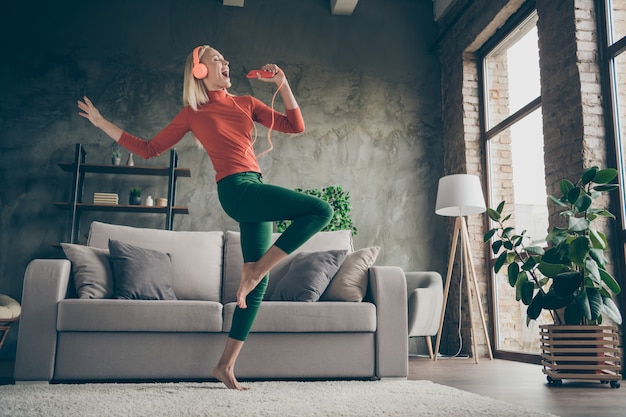 Full body low below angle view photo of pretty lady holding telephone listening song modern orange earphones dancing singing loudly wear casual outfit living room indoors