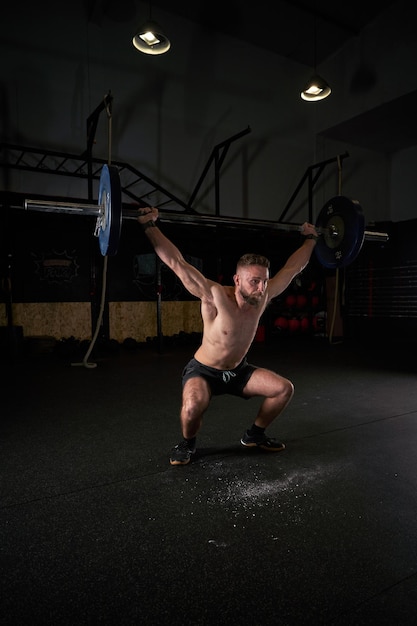 현대적이고 넓은 체육관에서 훈련하는 동안 근육질의 남성 보디빌더 전신이 쪼그리고 머리 위로 무거운 바벨을 들어올립니다.