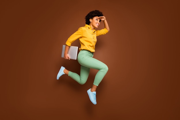 Full body photo of funky dark skin lady jump high hold notebook hurry lessons school see building close wear yellow shirt green pants footwear isolated brown color