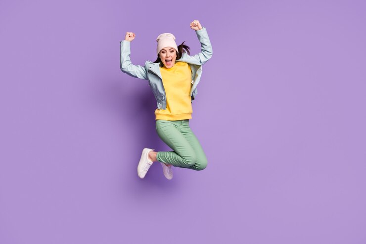 Photo full body photo of funky pretty lady stylish clothes jump high rejoicing champion screaming good mood wear casual hat jacket green pants footwear isolated purple color background
