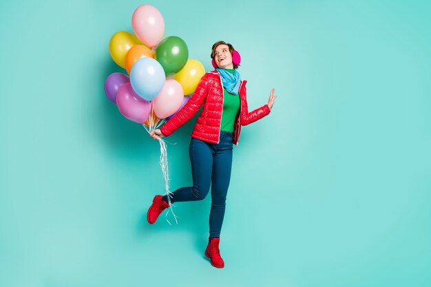 Foto di tutto il corpo di una signora agghiacciante divertente è venuta alla festa di compleanno portare molti palloncini colorati indossare sciarpa cappotto rosso casual cuffie auricolari rosa pantaloni scarpe isolato muro color verde acqua