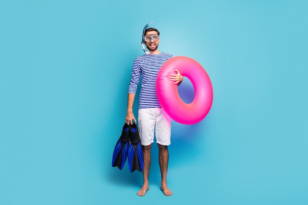 Full body photo of funny excited guy tourist swimmer hold underwater mask breathing tube flippers pink lifebuoy wear striped sailor shirt shorts isolated blue color