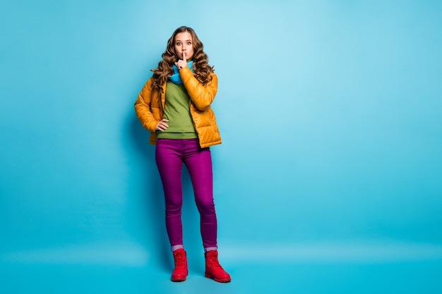 Full body photo of pretty curly lady holding finger on lips asking keep secret in privacy wear casual yellow overcoat scarf violet trousers red shoes isolated blue color wall