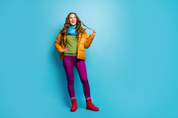 Full body photo of pretty lady enjoy spring sunny day look side empty space playing one curl wear casual yellow overcoat scarf violet pants red shoes isolated blue color wall