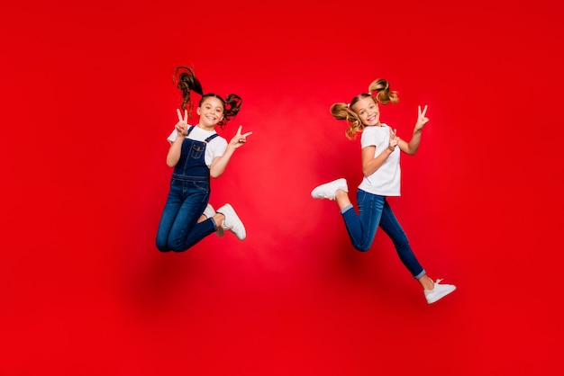 Фото всего тела двух позитивных светлых брюнеток с волосами, дети, девочки с хвостами, ждут рождественских праздников, прыгают, заставляют v-знаки носить современные повседневные футболки, изолированные на красном фоне