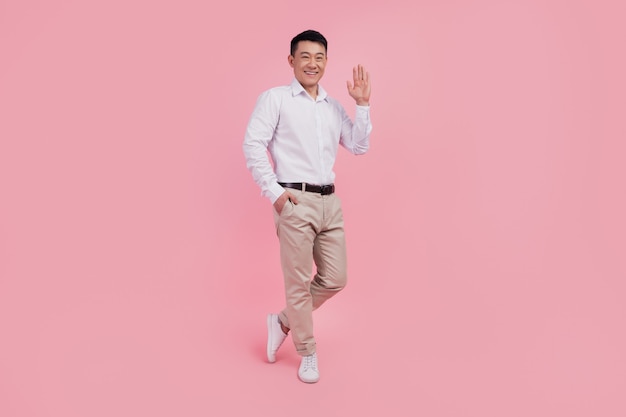 Full body photo of young asian man waving hand hello greeting friendly isolated over pink color background