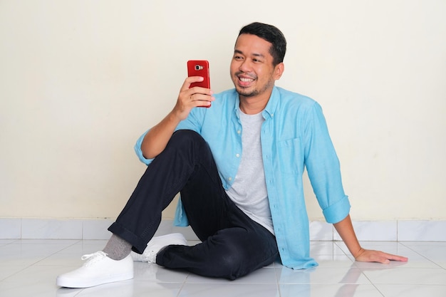 Full body portrait of adult Asian man sitting on the floor smiling when looking to his mobile phone