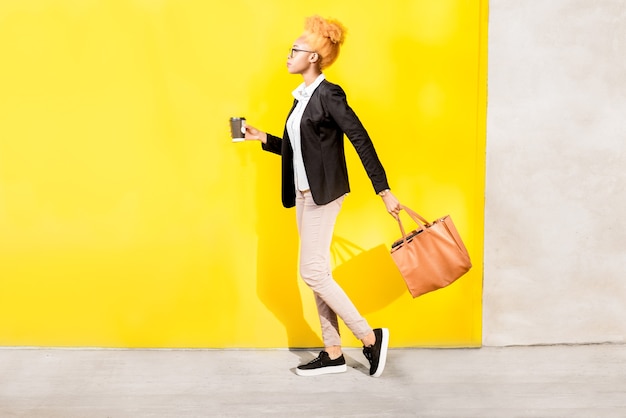 Full body portrait of an african business lady in casual suit walking with bag on the yellow wall background