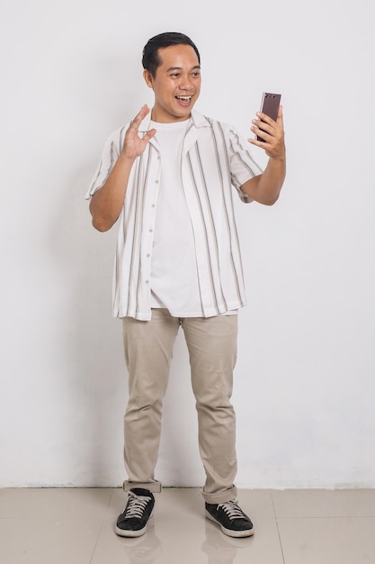 화상 통화를 하는 동안 휴대전화를 보며 행복한 인사 제스처를 하는 아시아 남성의 전신 초상화