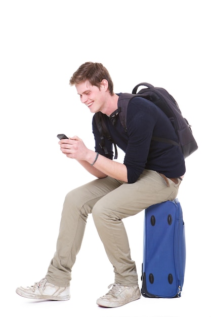 スーツケースの上に座って携帯電話を見ている幸せな若い男の全身ポートレート