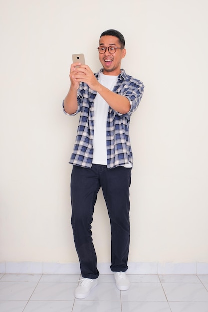 Photo full body portrait of a man looking to mobile phone that he hold with surprised expression