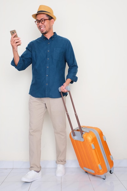 Full body portrait of a man pulling suitcase and looking to mobile phone with happy expression