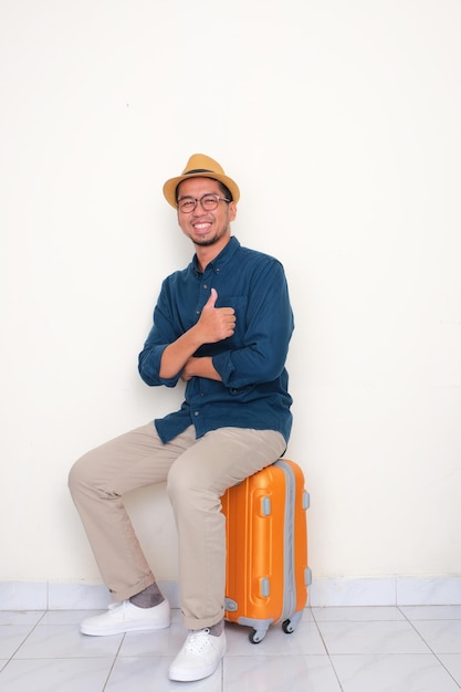 Full body portrait of a man smiling and give thumb up while sitting on suitcase