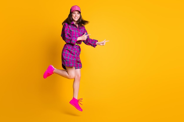 Full body portrait of pretty girl jump finger point empty space headwear plaid outfit isolated on yellow color background