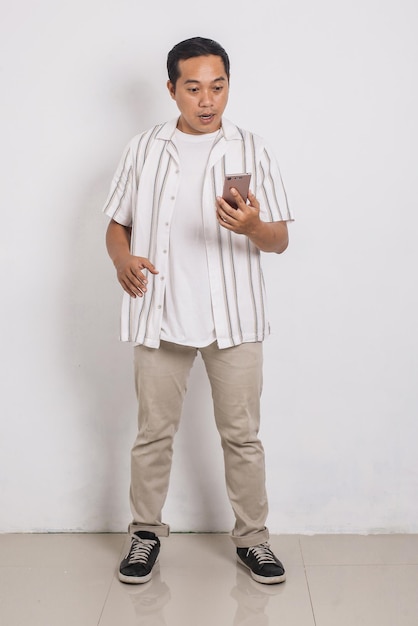 Full body portrait of surprised asian man pensive looking at cell phone on white background