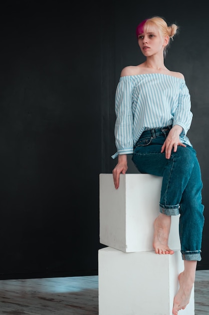 Full body portrait of young woman with white and pink hair\
sitting on white cube on black background