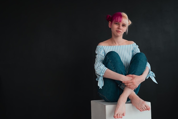 Full body portrait of young woman with white and pink hair
sitting on white cube on black background