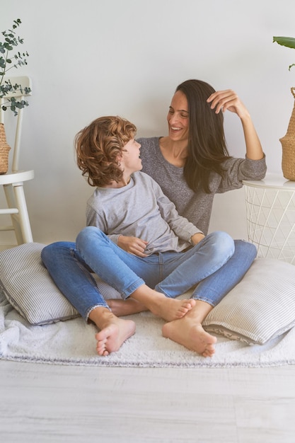 Full body of positive barefoot mother with kid looking at each other while sitting on blanket with cushions in living room