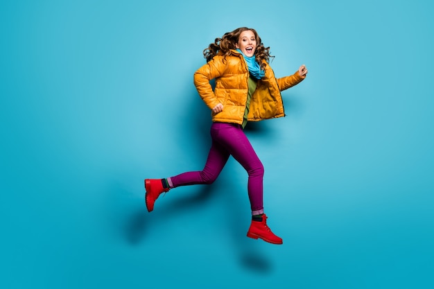 Full body profielfoto van mooie dame springen hoog haasten lage prijzen winkelen dragen casual gele overjas sjaal magenta broek rode schoenen geïsoleerde blauwe kleur muur