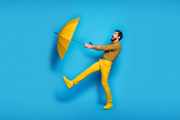 Full body profile side photo of scared frustrated anxious man travel windy weather catch flying bright parasol wear checkered trousers sneakers isolated over blue color