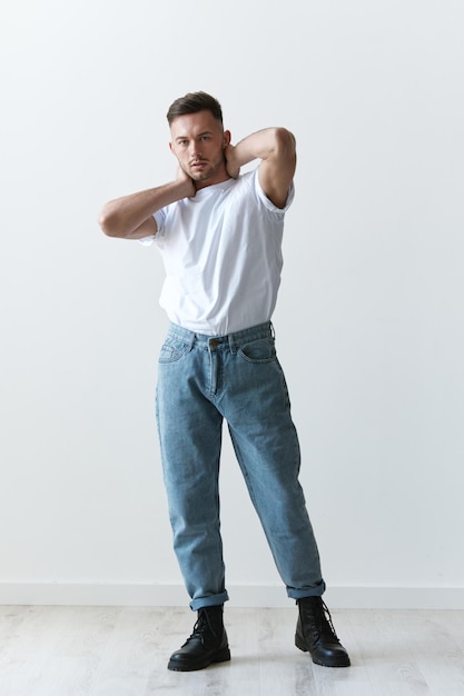 Full body shot of handsome serious tanned man guy in basic\
tshirt holds hand behind head posing on white background fashion\
style new collection offer copy space for ad modeling\
snapshots