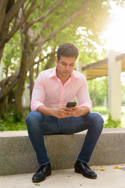 Full body shot of young handsome Indian businessman using phone and sitting at the park