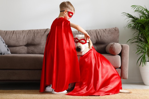 Full body side view of little boy and cute labrador retriever\
dog dressed in red superhero cloaks and glasses playing together at\
home