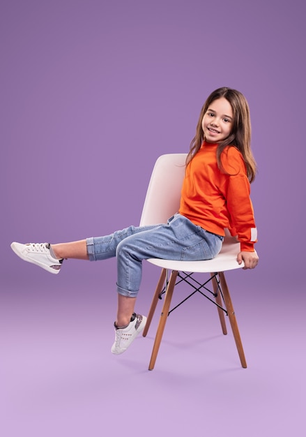 Full body side view of smiling little girl in orange sweatshirt and jeans with white sneakers sitting on chair in studio against violet background