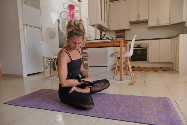 Full body of slim female in sportswear sitting in Padmasana on exercise mat while cuddling cat and doing yoga at home