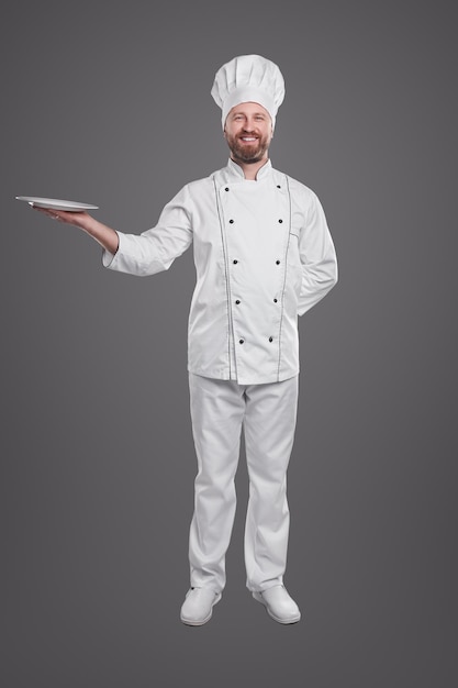 Full body of a smiling chef in uniform holding empty plate isolated against dark background