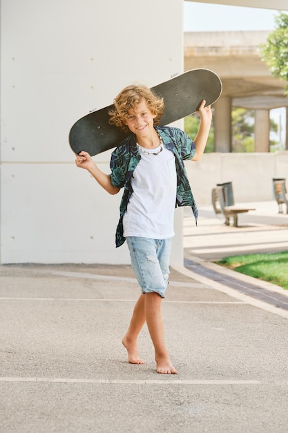 Full body of smiling child in casual clothes walking on city street with skateboard in hands and looking at camera on bright summer day